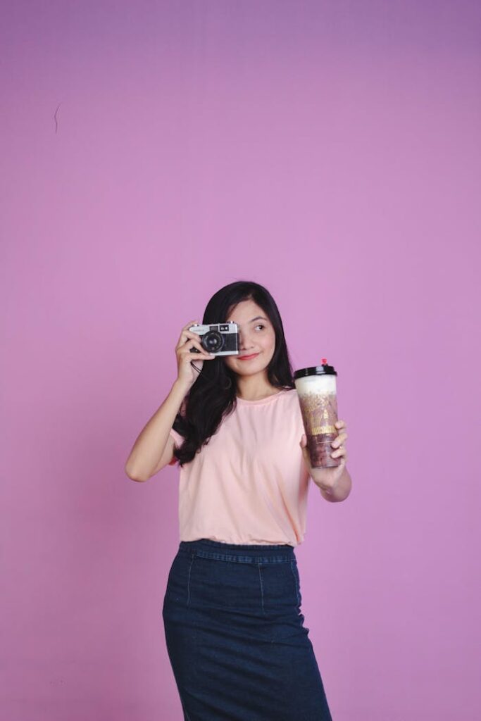 pexels-photo-6368572 Smiling young woman holding a camera and bubble tea against a pink studio backdrop.